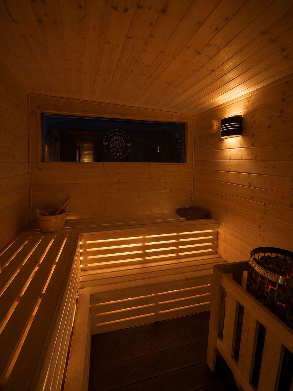 Saunaraum im Wellness Cube bei gedimmtem Licht: beleuchtete Bänke, Wandleuchte, Ofen mit Steinen.